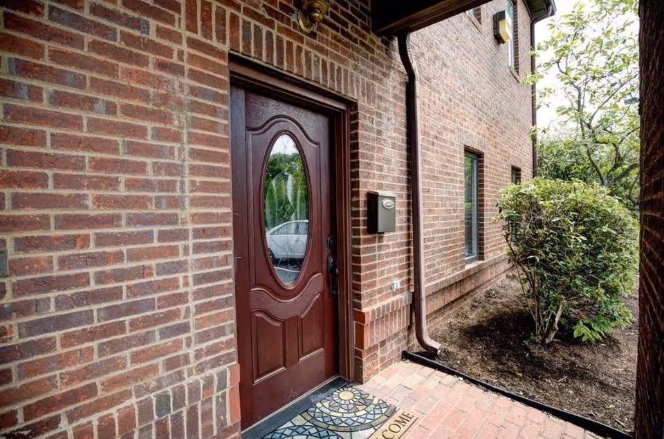 Brick exterior entrance with a maroon door, window, a welcome mat, and surrounding shrubs.