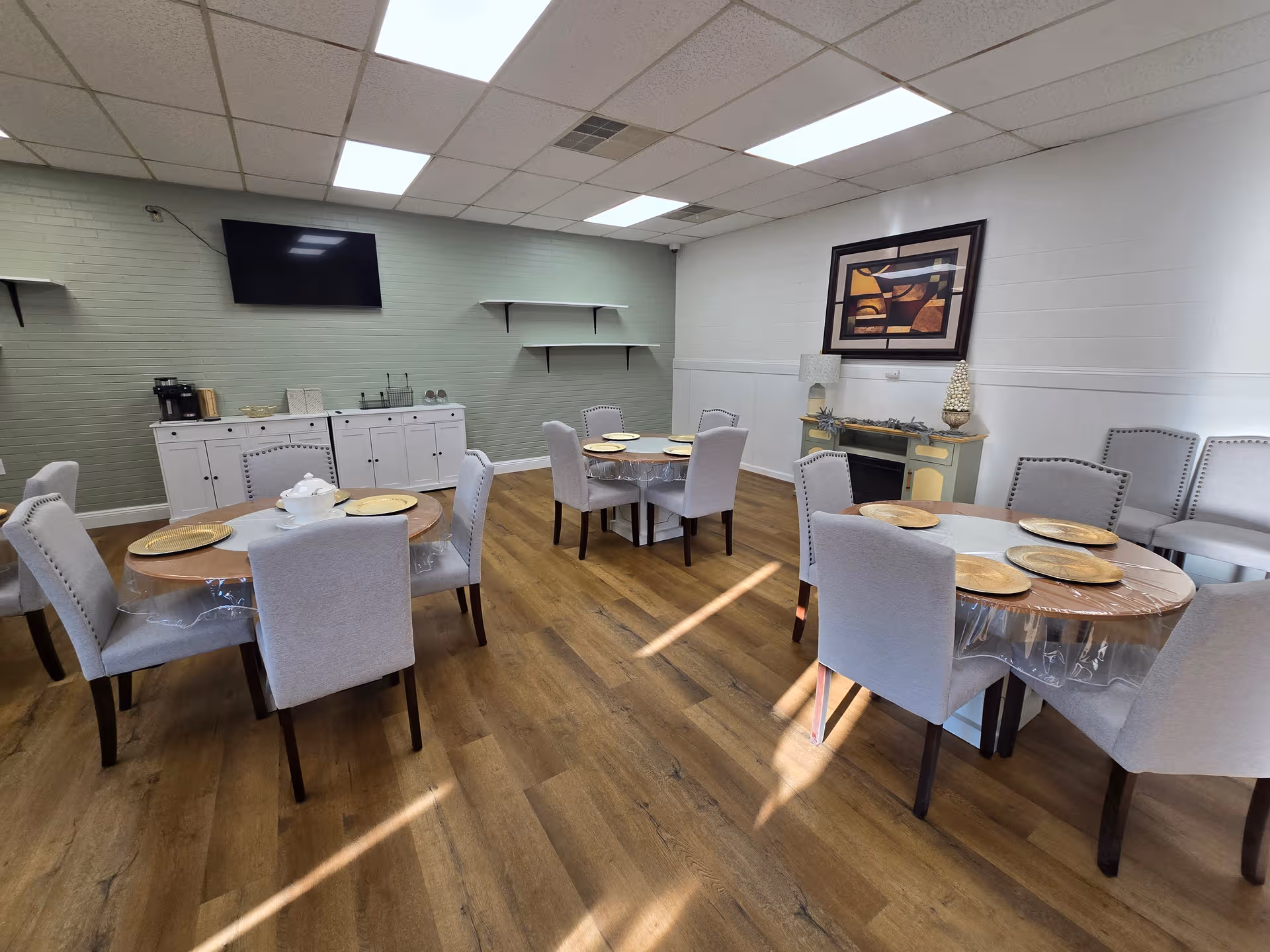 A dining room with round tables, upholstered chairs, and a kitchenette area along the wall.