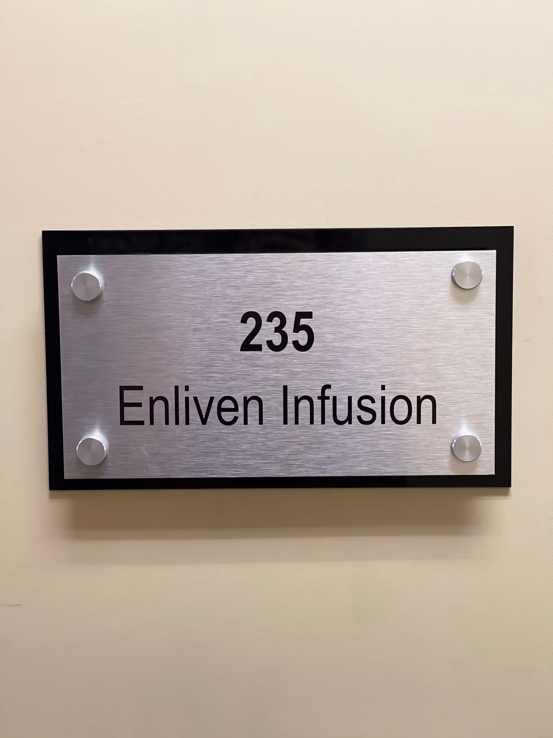 Metal wall plaque reading '235 Enliven Infusion' mounted in a hallway.
