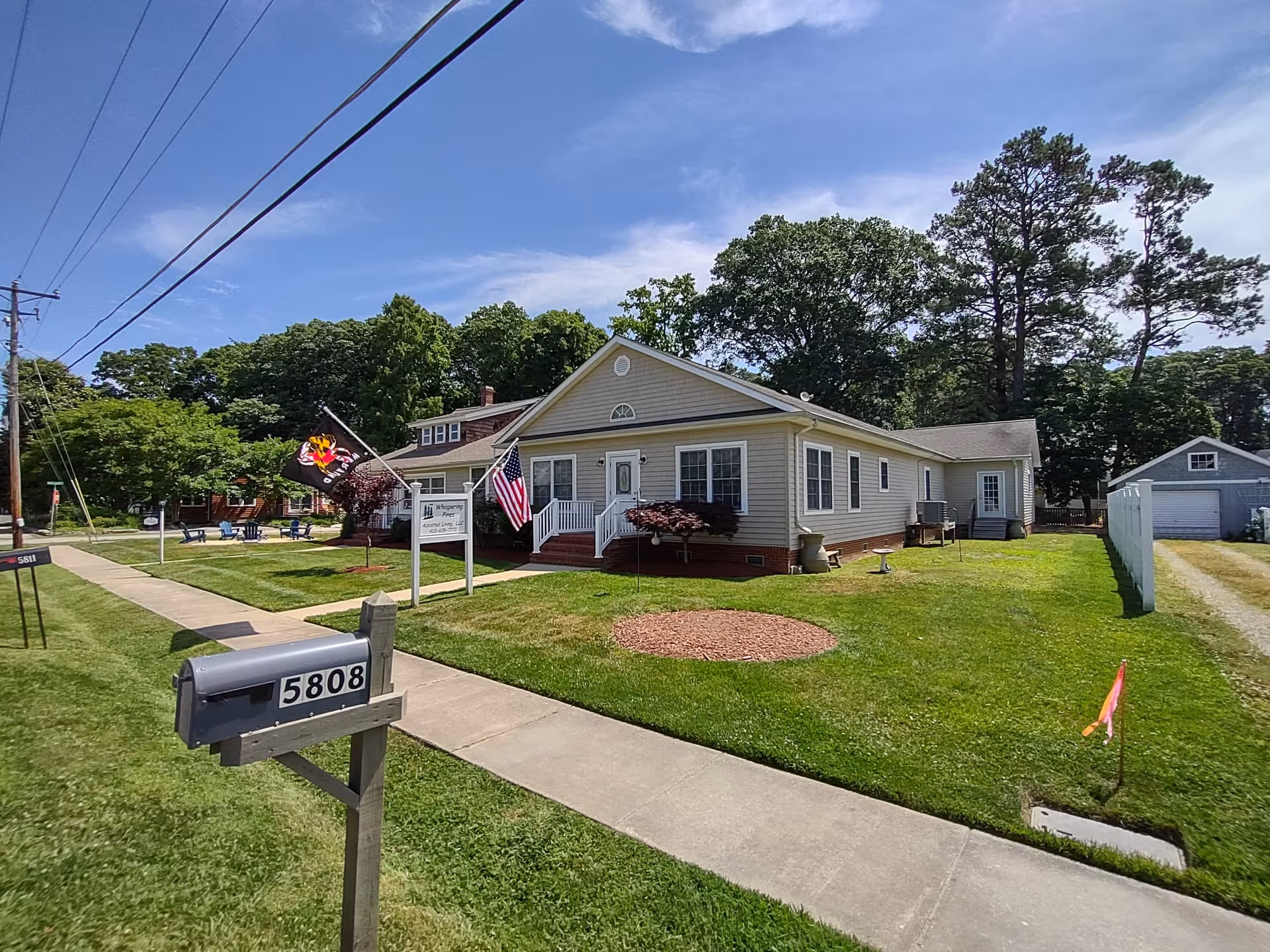 Exterior view of a single-story residential building with beige siding and white trim, identified as Whispering Pines Assisted Living Facility. The building has a small porch with white railings, two flags displayed near the entrance, and a well-maintained lawn with a circular mulch bed. A mailbox with the number 5808 is visible in the foreground, along with a sidewalk and a driveway leading to a detached garage. Trees and a clear blue sky are in the background.