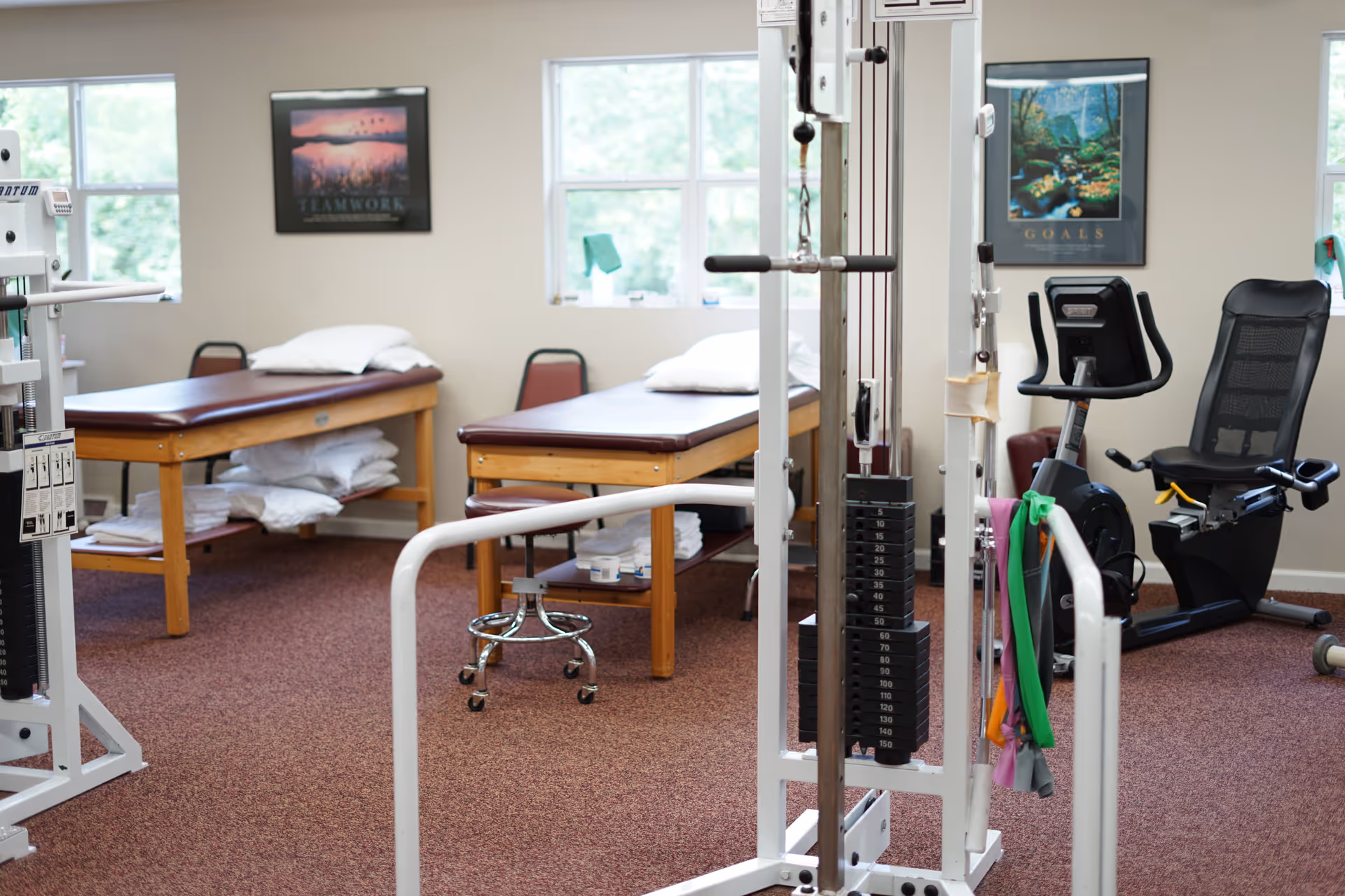 Physical therapy room with treatment beds, exercise machines, and mirrors; windows in background.