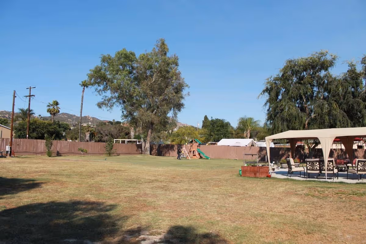 Large grassy outdoor yard with trees, a wooden fence, a covered seating area and a small playground under a clear blue sky.