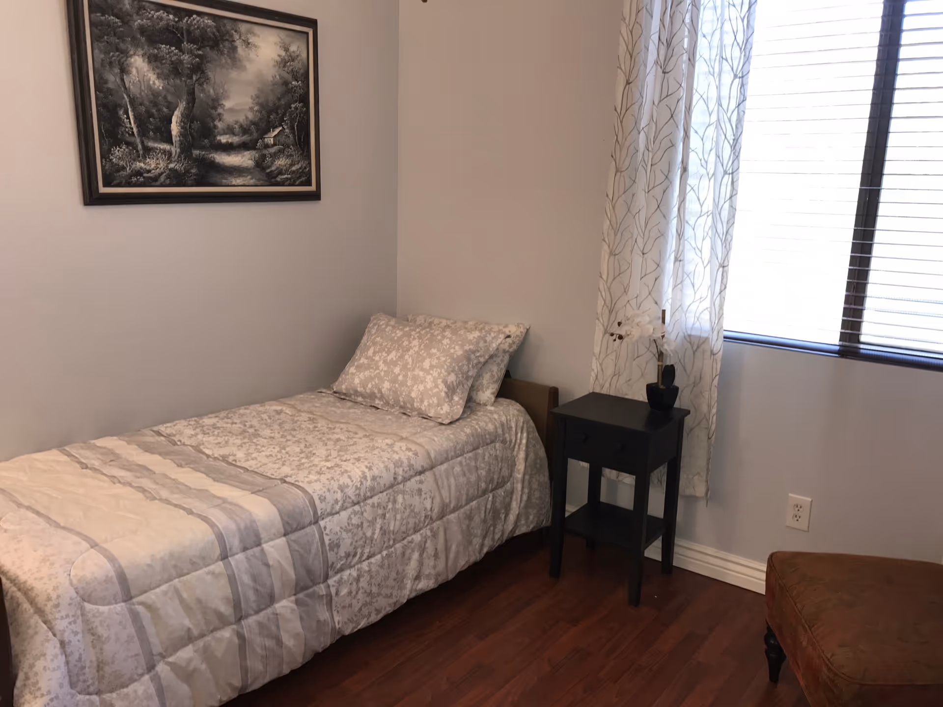 A small bedroom with a single bed covered in a light-colored patterned comforter and matching pillow. Next to the bed is a black nightstand with a small potted plant on top. A window with sheer white curtains and blinds is on the right wall. A framed black and white landscape painting hangs on the wall above the bed. The floor is wooden, and part of a brown upholstered chair is visible in the corner.