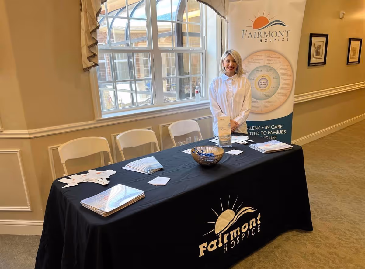 Smiling staff member stands behind a promotional table with Fairmont Hospice banner indoors.