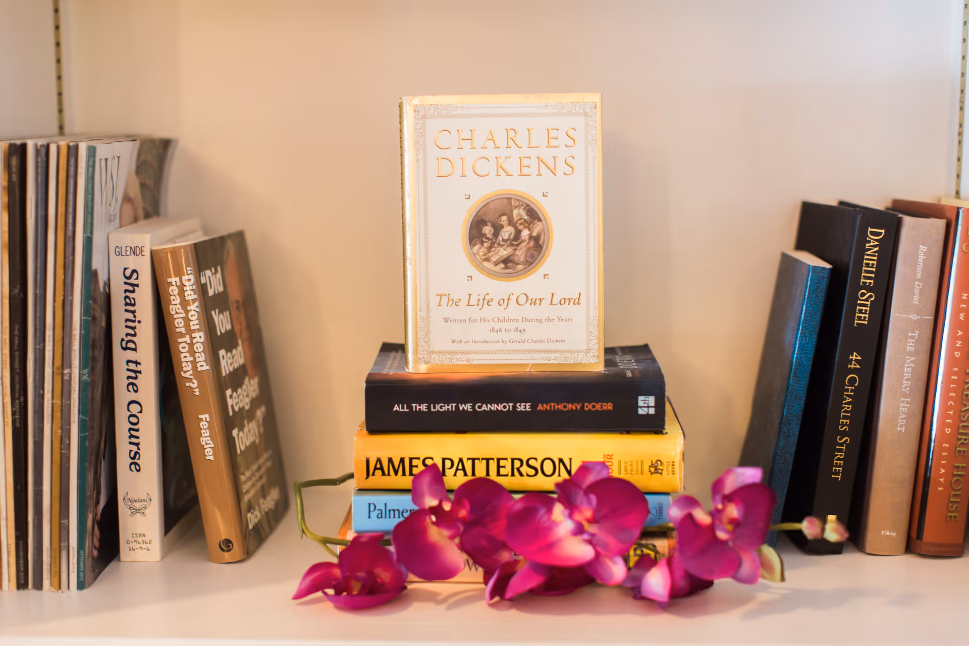 A close-up view of a bookshelf with various books arranged vertically and horizontally. Prominently displayed in the center is a book titled 'Charles Dickens: The Life of Our Lord' standing upright on top of a stack of three books. In front of the stack is a purple orchid flower lying horizontally. Other visible book titles include 'Did You Read Feagler Today?', '44 Charles Street' by Danielle Steel, and 'Sharing the Course'.