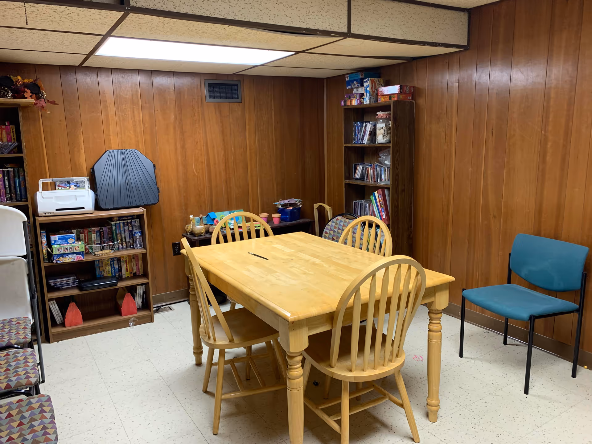 Wood-paneled activity room with a large table, chairs, and shelving.