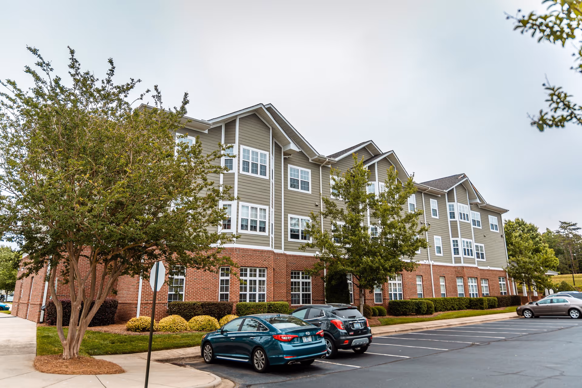 Exterior view of a three-story senior living facility building with a brick lower level and gray siding upper levels. Several windows are visible, along with a parking lot in front containing a few parked cars. Trees and shrubs are planted around the building and along the sidewalk.
