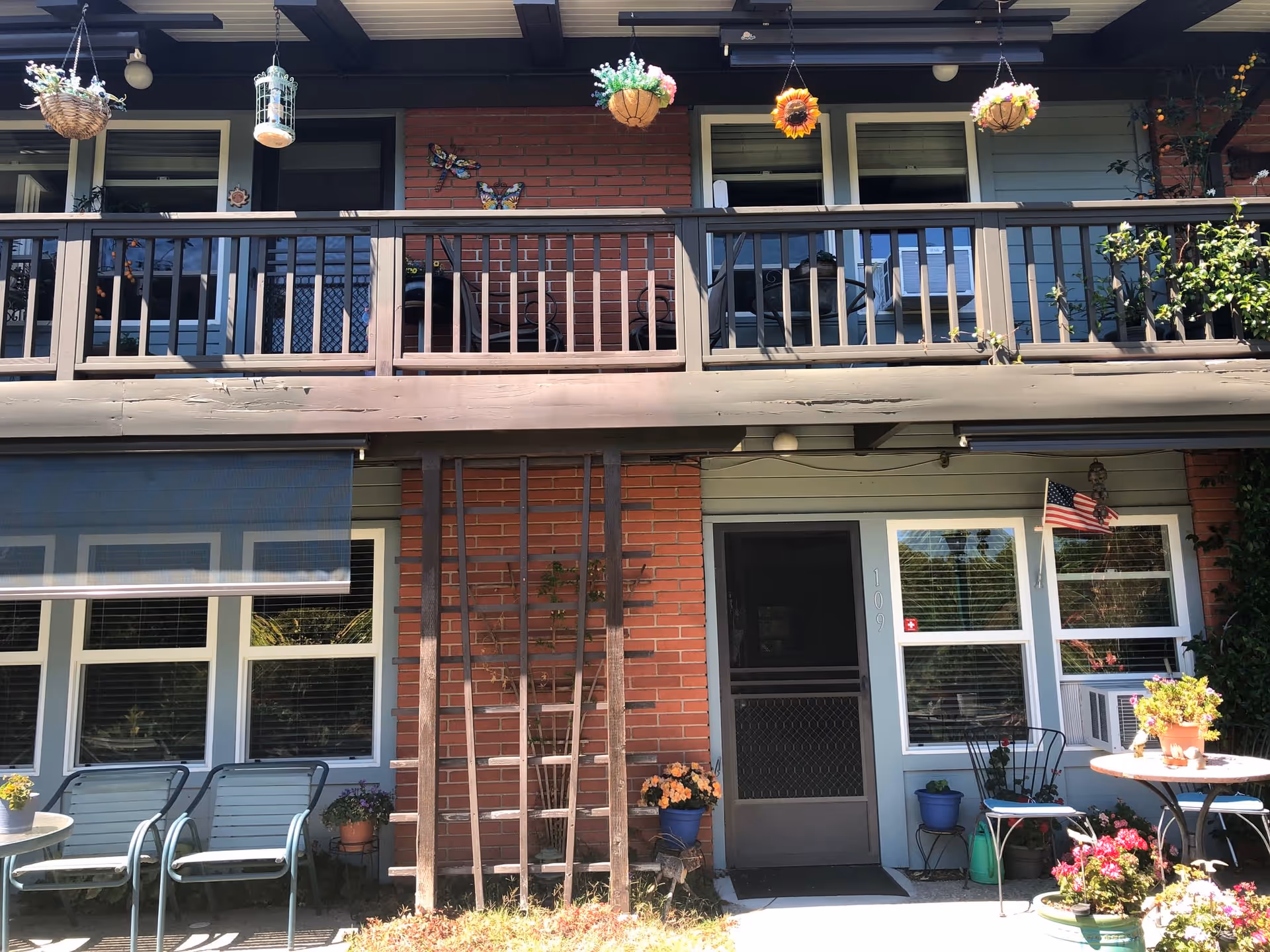 Exterior view of a two-story residential building with a balcony on the upper floor. The building features a combination of brick and light blue siding. Several hanging flower baskets and potted plants decorate the balcony and ground level. There are chairs and a small table with flowers on the patio area. An American flag is displayed near a window with an air conditioning unit.