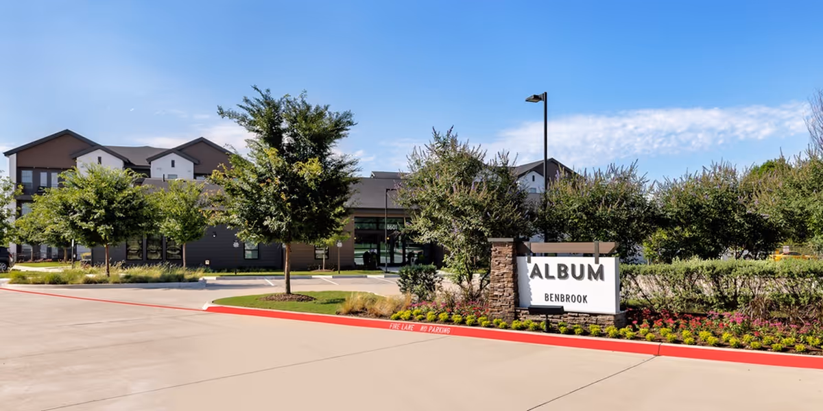 Exterior view of Album Benbrook senior living facility with a clear blue sky, landscaped greenery, and a sign displaying the facility's name near the entrance driveway.
