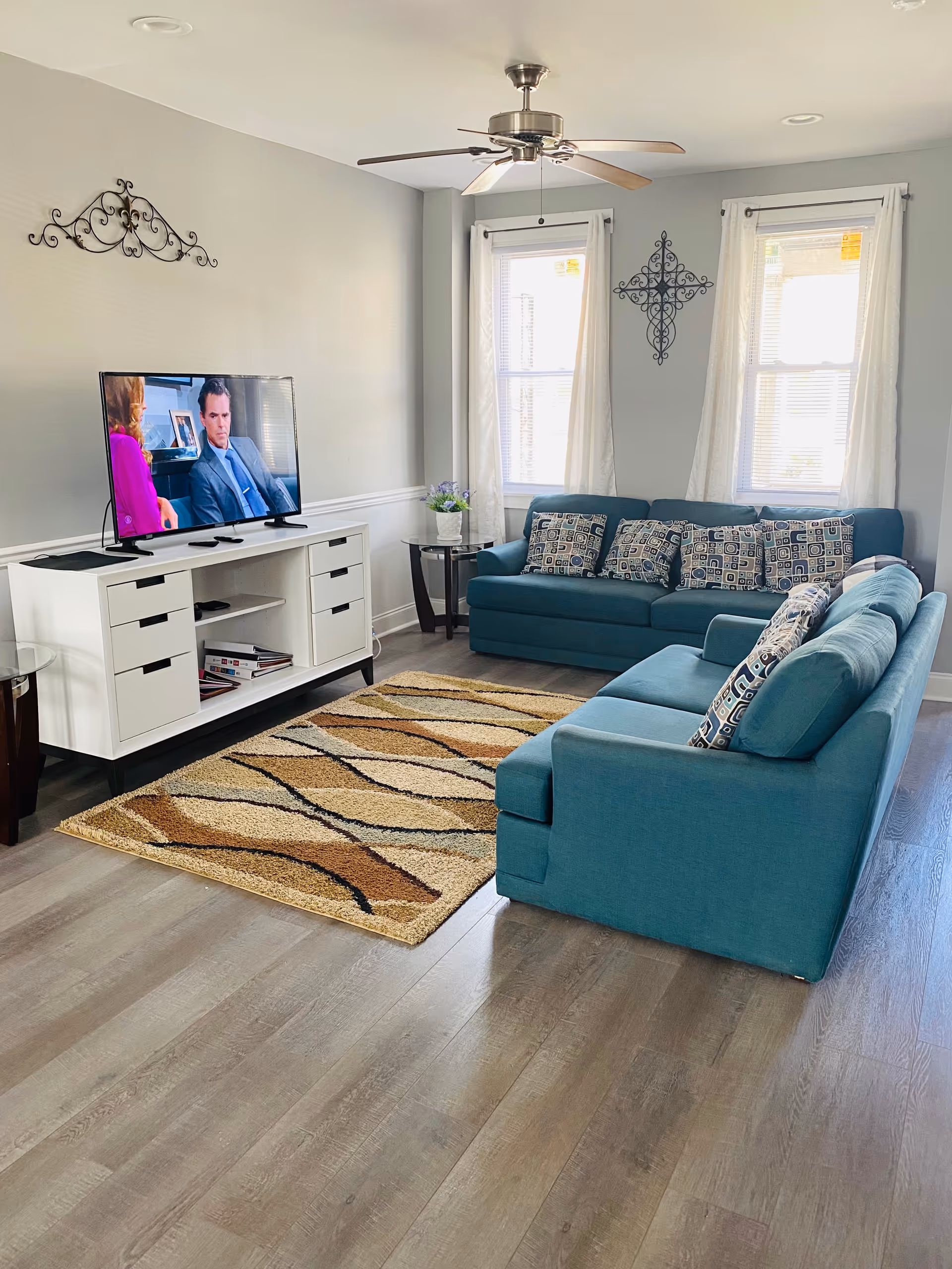 Bright living room with teal sofas, rug, TV, and windows with sheer curtains.