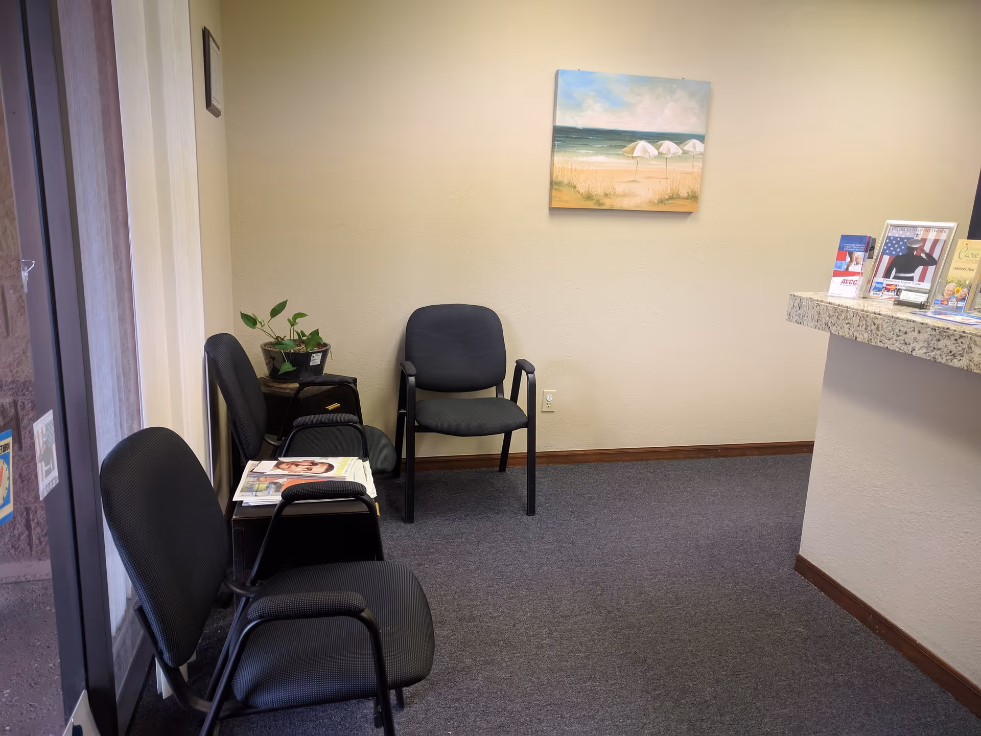 Waiting area with black chairs, magazines, a plant, and a beach-themed painting.