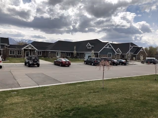 Exterior view of a single-story senior living facility building with a dark roof and multiple windows. Several cars are parked in the parking lot in front of the building. The sky is partly cloudy and there is a grassy area with small trees in the foreground.