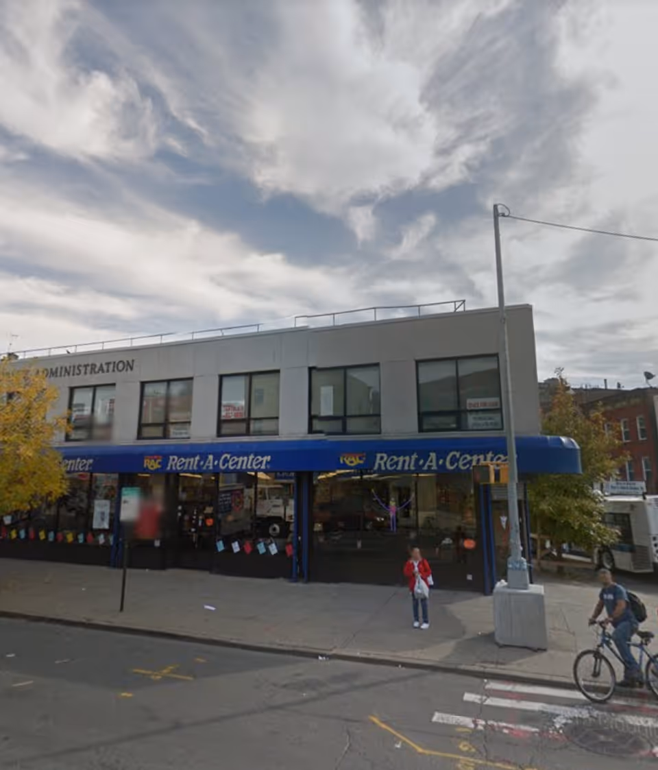 Two-story storefront building with blue Rent-A-Center sign on a city street, pedestrians nearby.