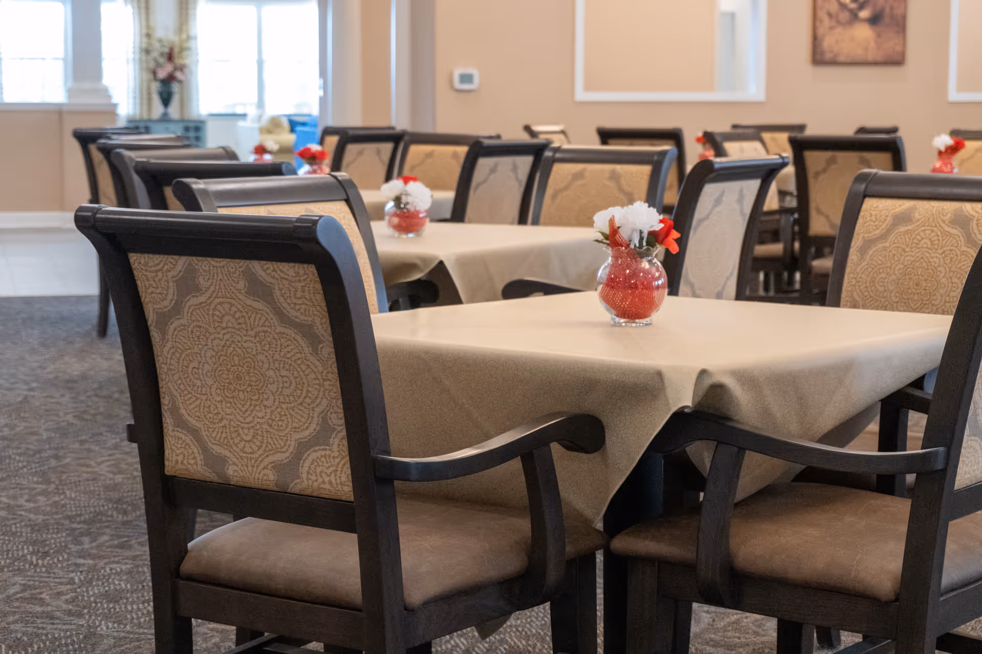 A dining room with multiple tables covered in beige tablecloths, each table decorated with a small glass vase containing red and white flowers. The chairs have dark wooden frames with patterned beige upholstery. The room has a carpeted floor and light-colored walls with framed artwork.