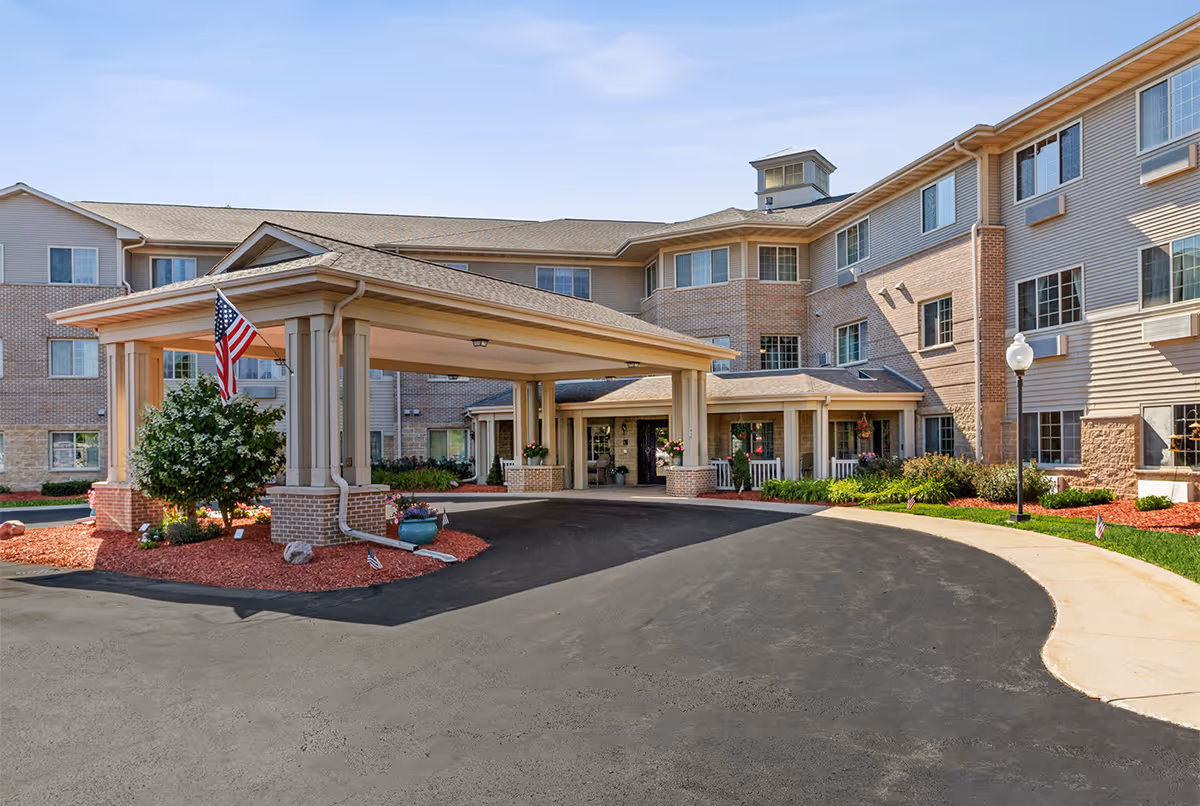 Front view of a multi-story senior living facility entrance with circular driveway and flag.