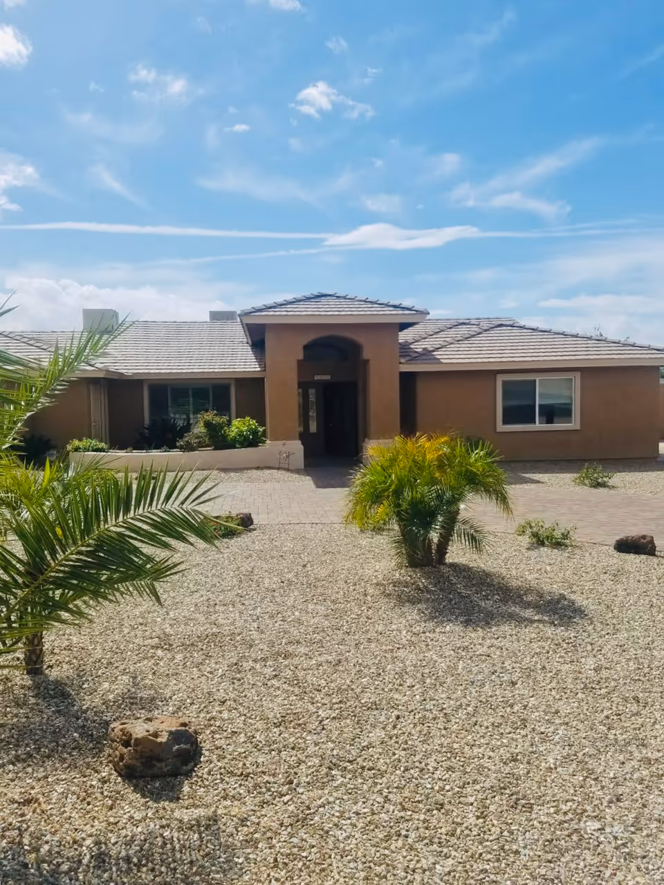 Front exterior view of a single-story building with a tiled roof and a central entrance. The yard is covered with gravel and has a few small palm trees and plants. The sky is clear with some clouds.