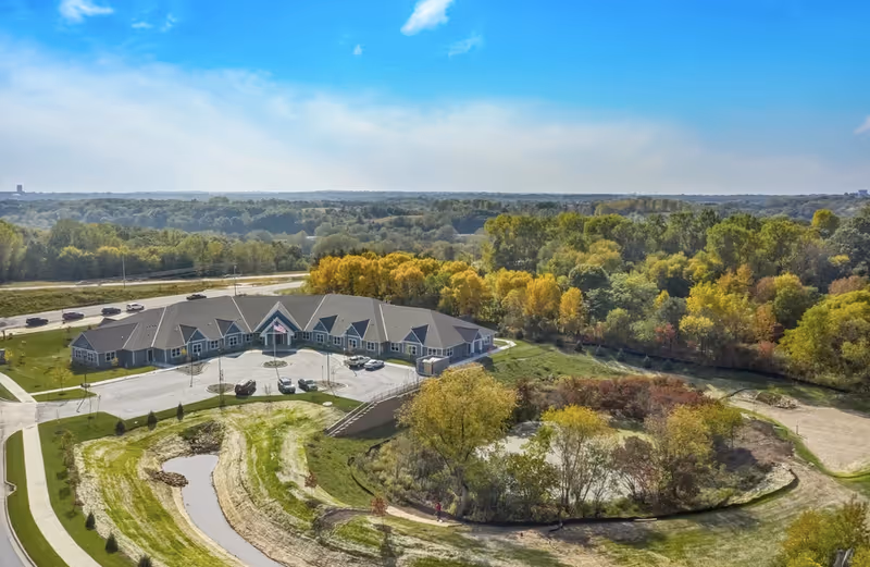 Aerial view of a single-story senior living facility surrounded by trees, lawns, a parking area, and a small pond under a blue sky.