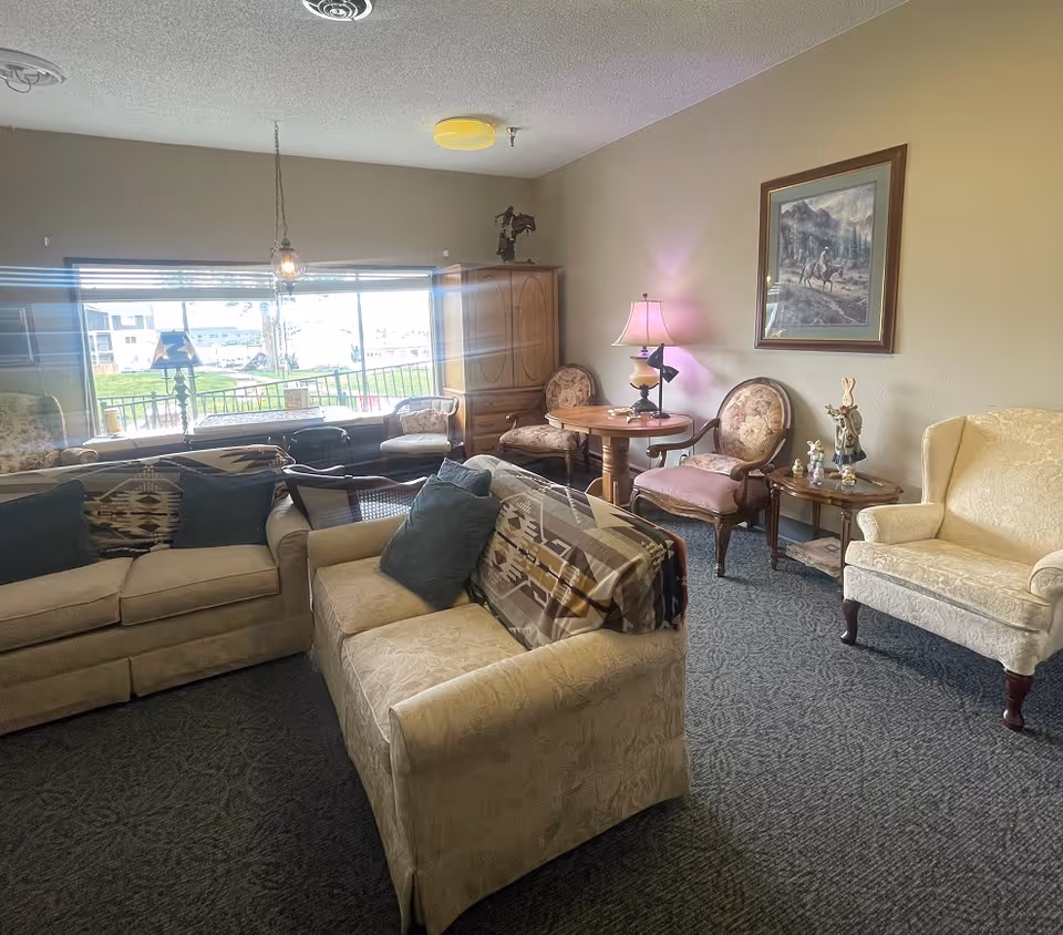 Cozy retirement lounge with beige sofas, chairs, a round table, and a large front window.