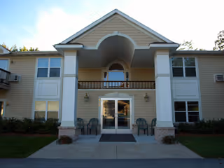 Front entrance of a two-story beige assisted living building with a covered portico, double glass doors, porch chairs, and multiple windows.