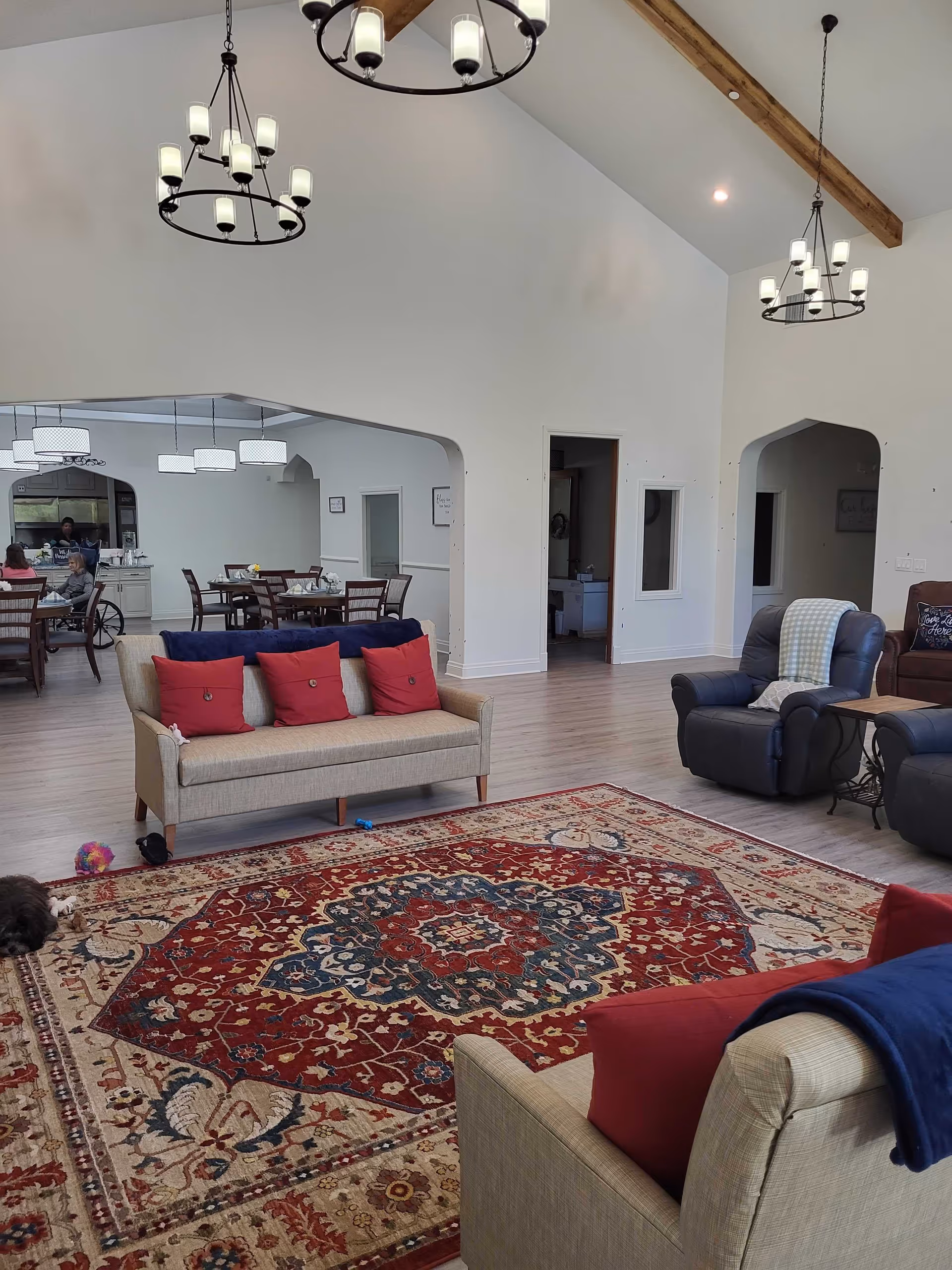 A spacious living room area with high vaulted ceilings and wooden beams. The room features two beige sofas with red pillows, two dark blue recliners, and a large ornate red and beige patterned rug. In the background, there is a dining area with tables and chairs, and a few people seated. The walls are white, and there are three circular chandeliers hanging from the ceiling.