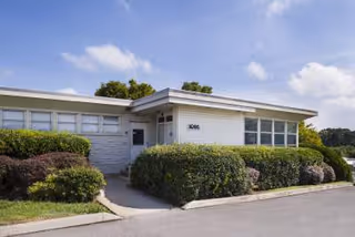 Single-story white building with large windows, trimmed hedges, and a paved entrance.