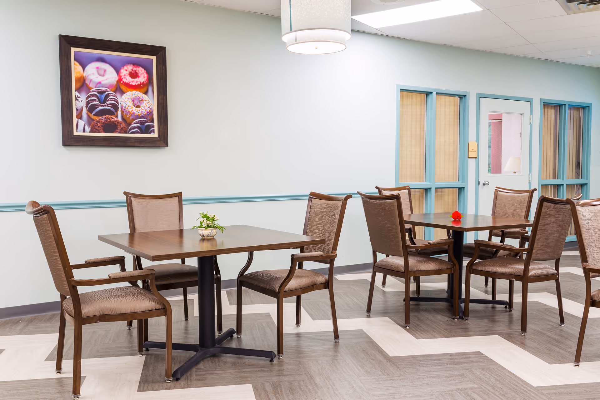 A dining area with two square wooden tables, each surrounded by four cushioned chairs with wooden frames. The floor has a zigzag pattern with light and dark wood tones. On the wall, there is a framed picture of colorful donuts. The walls are light-colored with a teal chair rail and window frames. A hanging light fixture is visible above the tables.