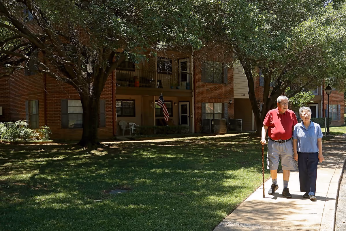 Two older adults walk along a shaded sidewalk in front of a brick apartment building with trees and an American flag.