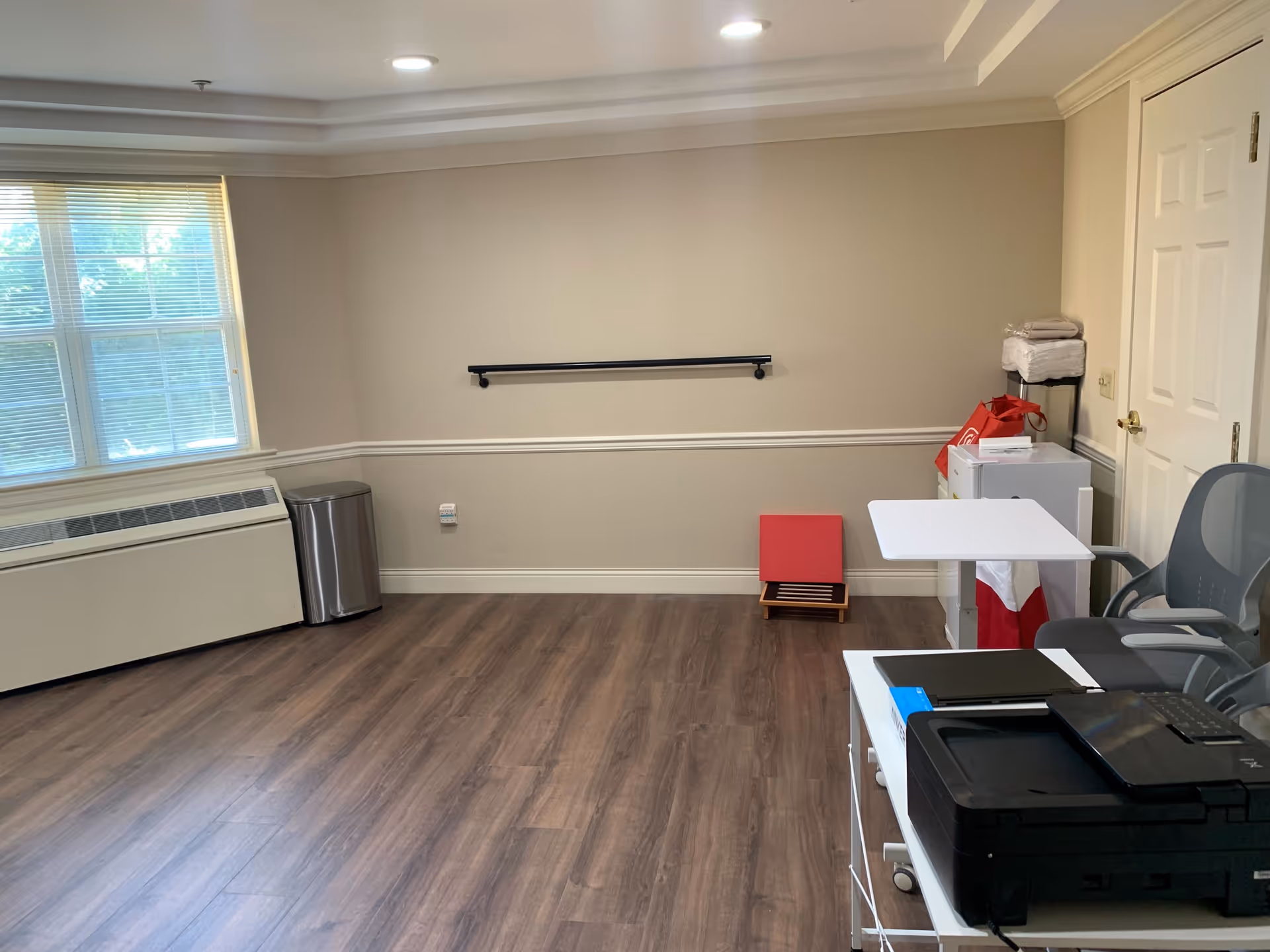 Interior senior facility room with a desk, printer, window, and wall-mounted handrail.