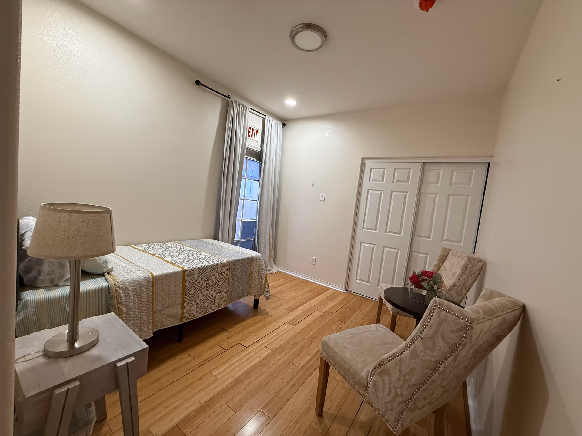 A small furnished bedroom with bed, nightstand, chairs, and a sliding closet in a retirement facility.