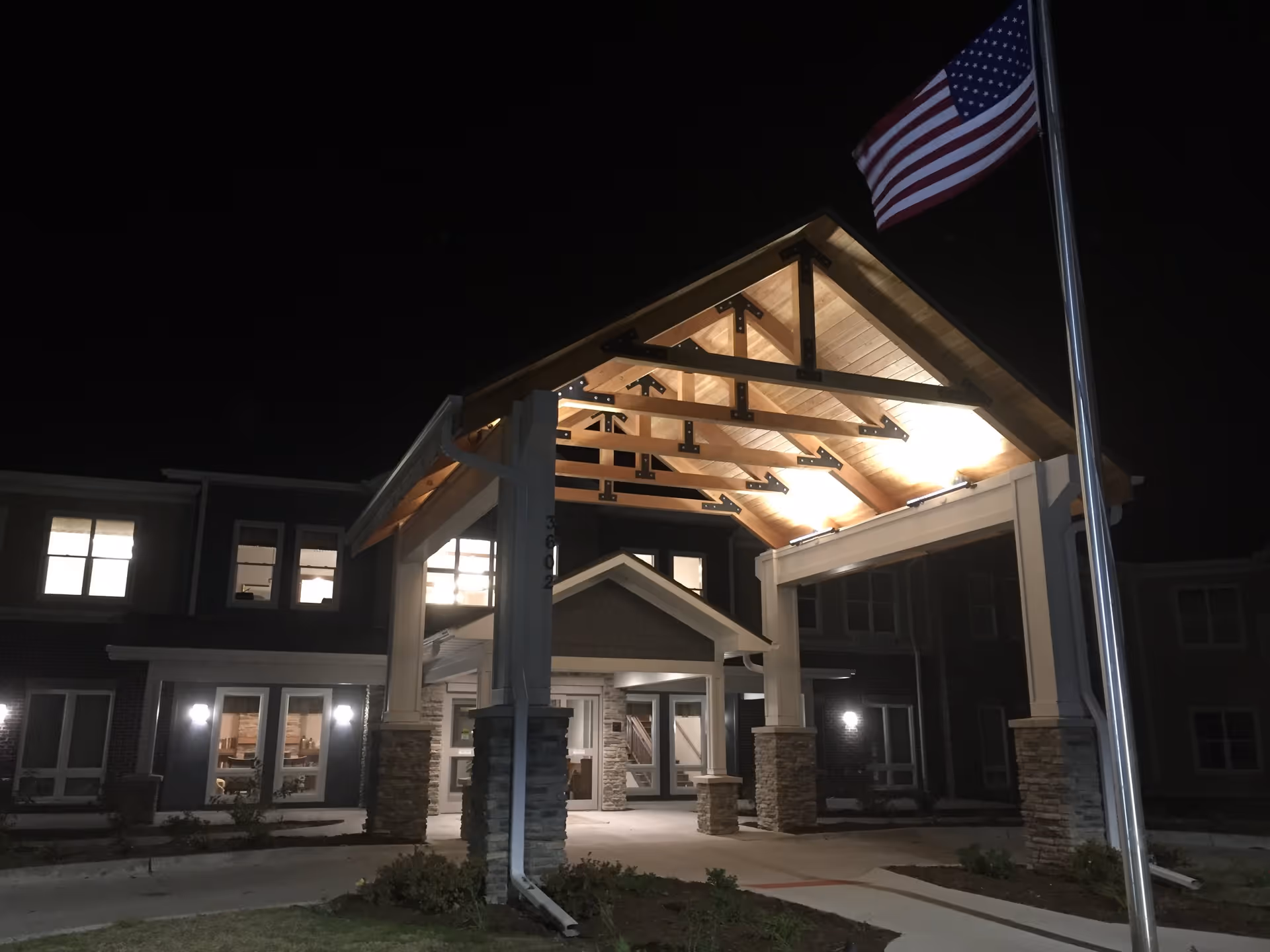 Night view of the entrance to a senior living facility with a well-lit wooden canopy supported by stone pillars, an American flag on a flagpole to the right, and the building's windows illuminated in the background.