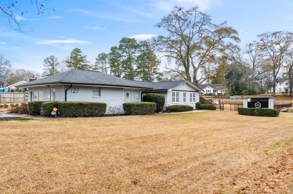 Single-story light-gray facility building with manicured hedges, a gate, and a wide grassy yard.