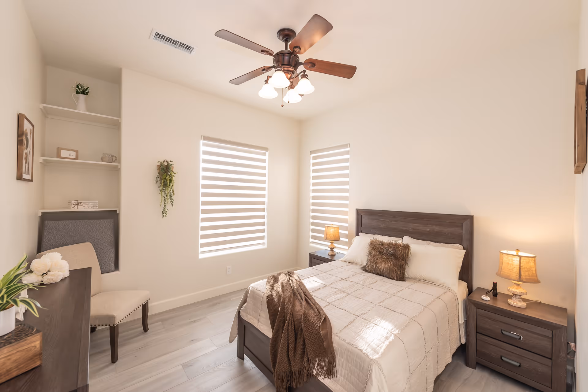 A bright and cozy bedroom with a wooden bed frame, beige bedding, and a brown throw blanket. The room features two windows with striped blinds, two bedside tables each with a lamp, a ceiling fan with lights, a beige upholstered chair, built-in shelves with decorative items, and light-colored wooden flooring.
