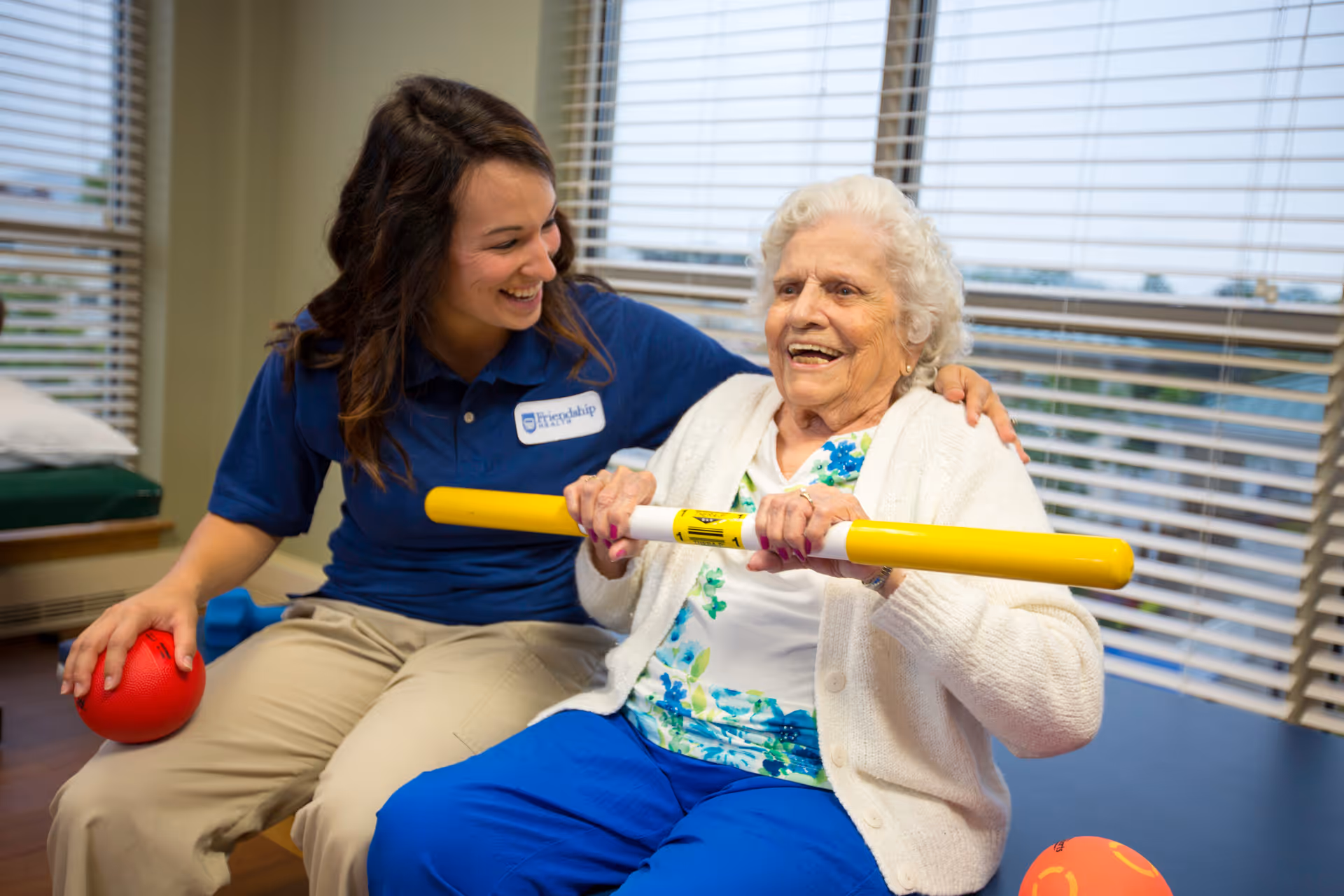 An elderly woman sitting and holding a yellow exercise bar, smiling, with a younger female caregiver in a blue Friendship Health & Rehab Center North shirt sitting beside her, also smiling and holding a red ball. They are in a room with large windows and blinds.