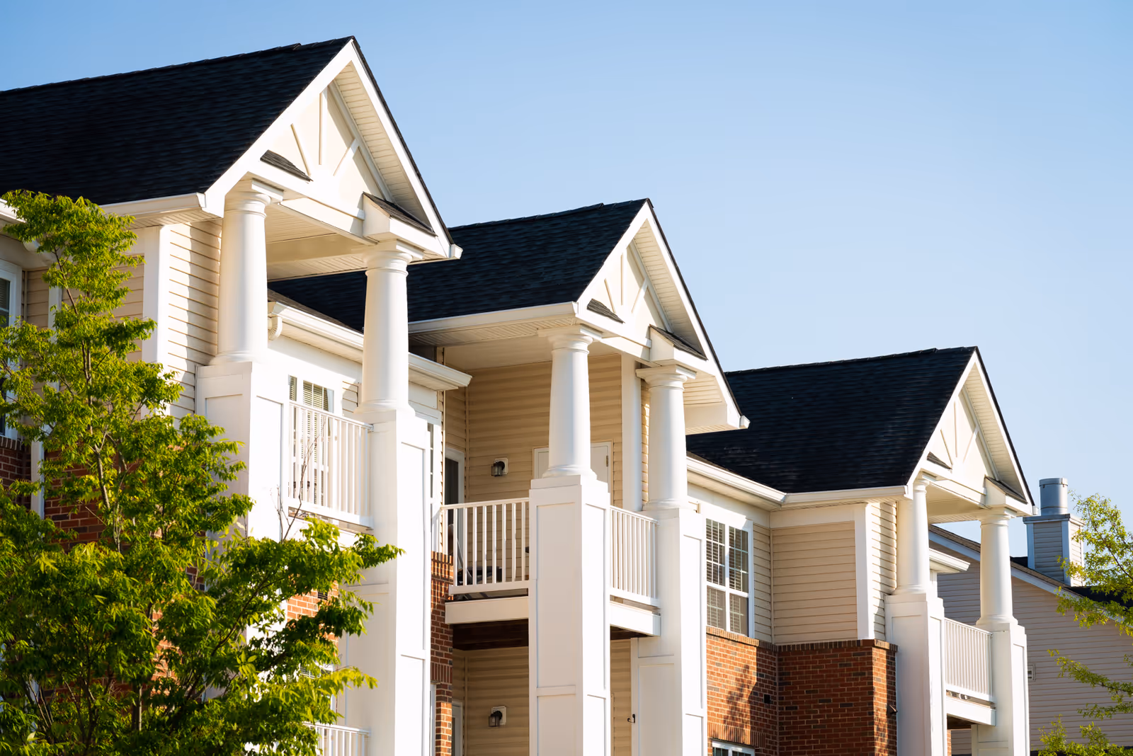 Exterior view of a residential building with beige siding, brick accents, white columns, and balconies under a clear blue sky with some green trees in the foreground.