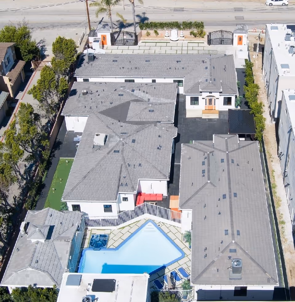 Aerial view of Oasis Homes facility showing multiple connected buildings with gray roofs, a fenced swimming pool with lounge chairs, a small green area with trees, and a gated entrance facing a street.