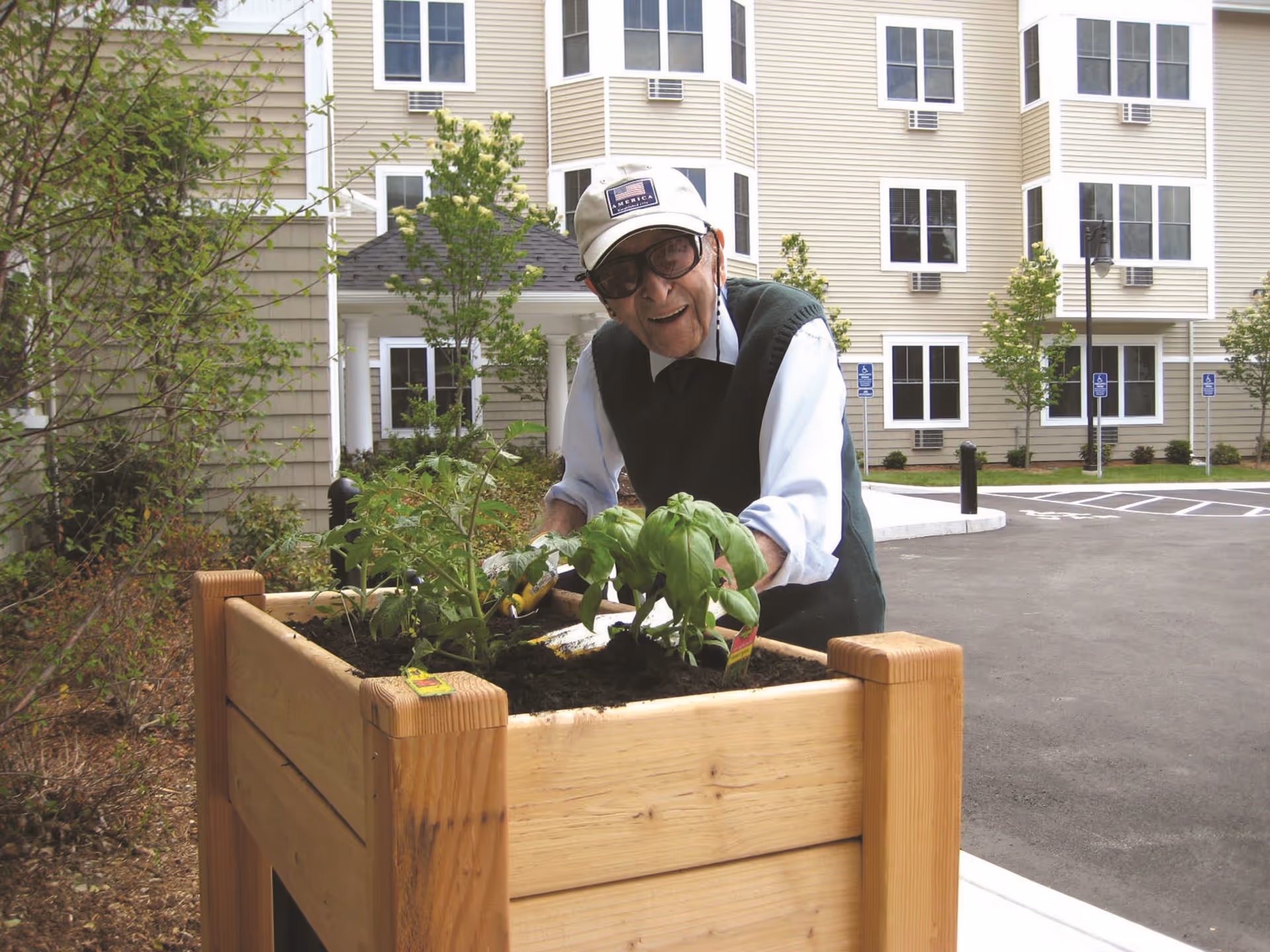 An elderly man wearing a white cap, glasses, and a dark green vest is gardening in a raised wooden planter box filled with soil and green plants. Behind him is a beige multi-story building with many windows and a parking lot with handicap parking signs.