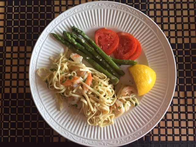 A white plate with a serving of shrimp pasta, steamed asparagus, two slices of tomato, and a lemon wedge on a woven placemat.
