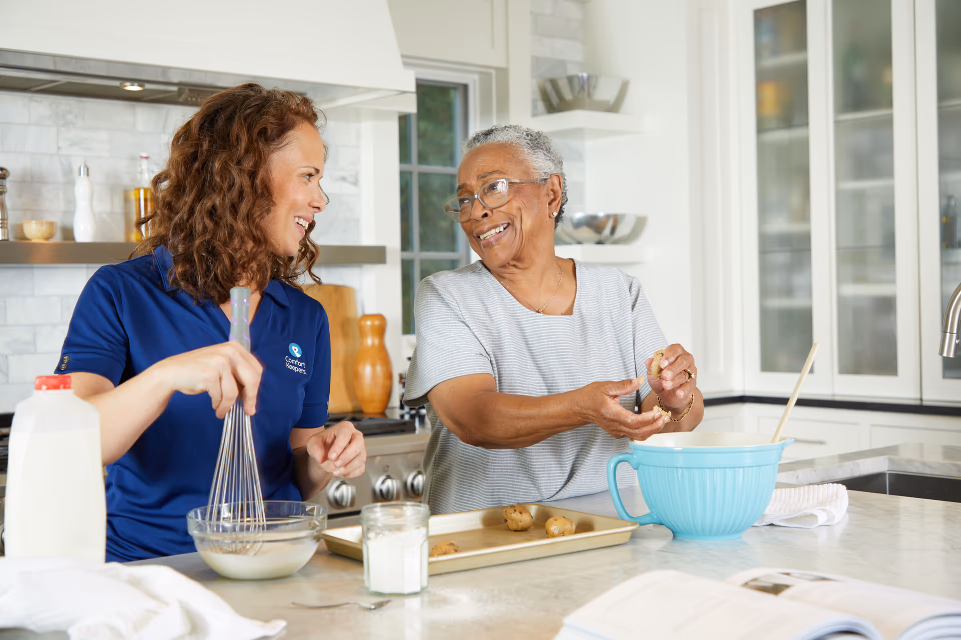 Caregiver in a blue uniform chats with an elderly woman while cooking in a bright kitchen.