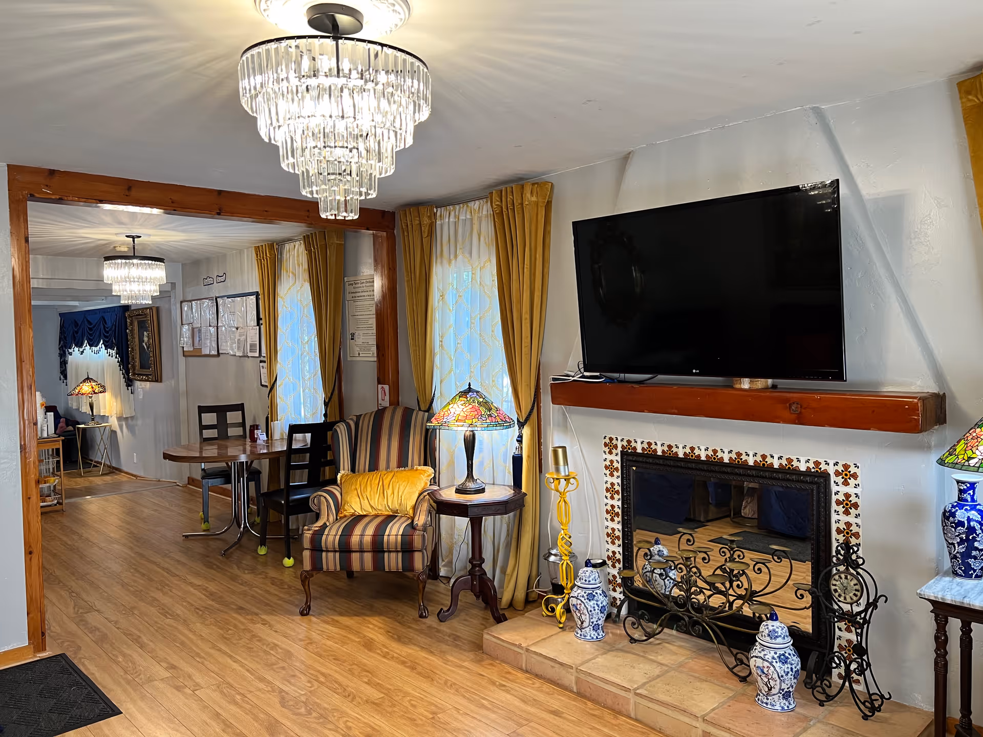 A cozy living room area in Fallbrook Assisted Living featuring a mounted flat-screen TV above a decorative fireplace with blue and white ceramic vases. There is a striped armchair with a yellow pillow next to a small wooden side table with a stained glass lamp. The room has wooden flooring, yellow curtains, and a crystal chandelier. In the background, there is a dining table with chairs and another stained glass lamp on a side table.