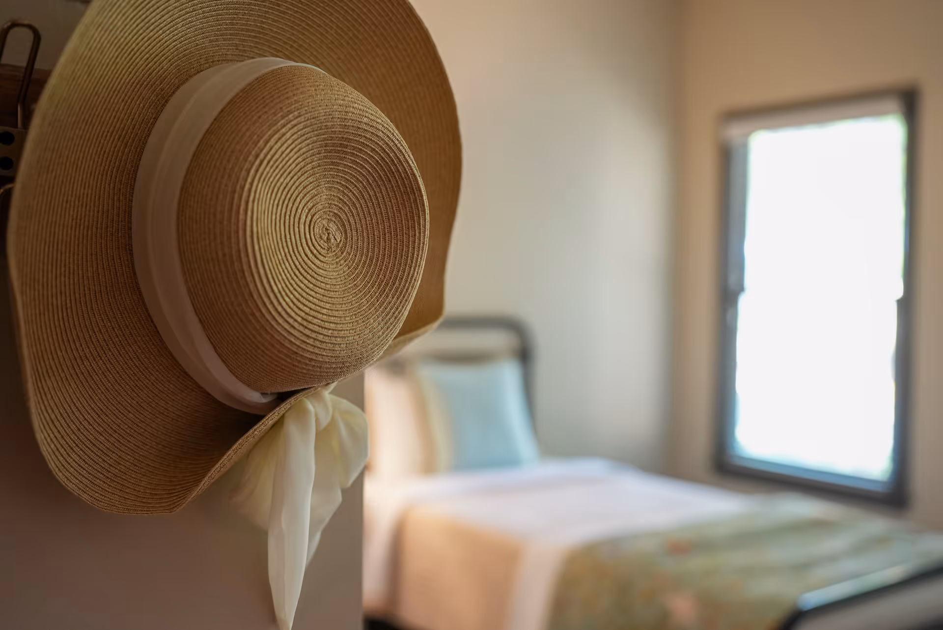 A straw sun hat hangs on a hook near a neatly made bedroom bed.