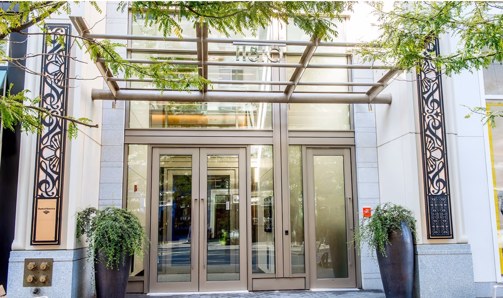 Modern building entrance with glass doors, metal frame, and potted greenery on both sides.