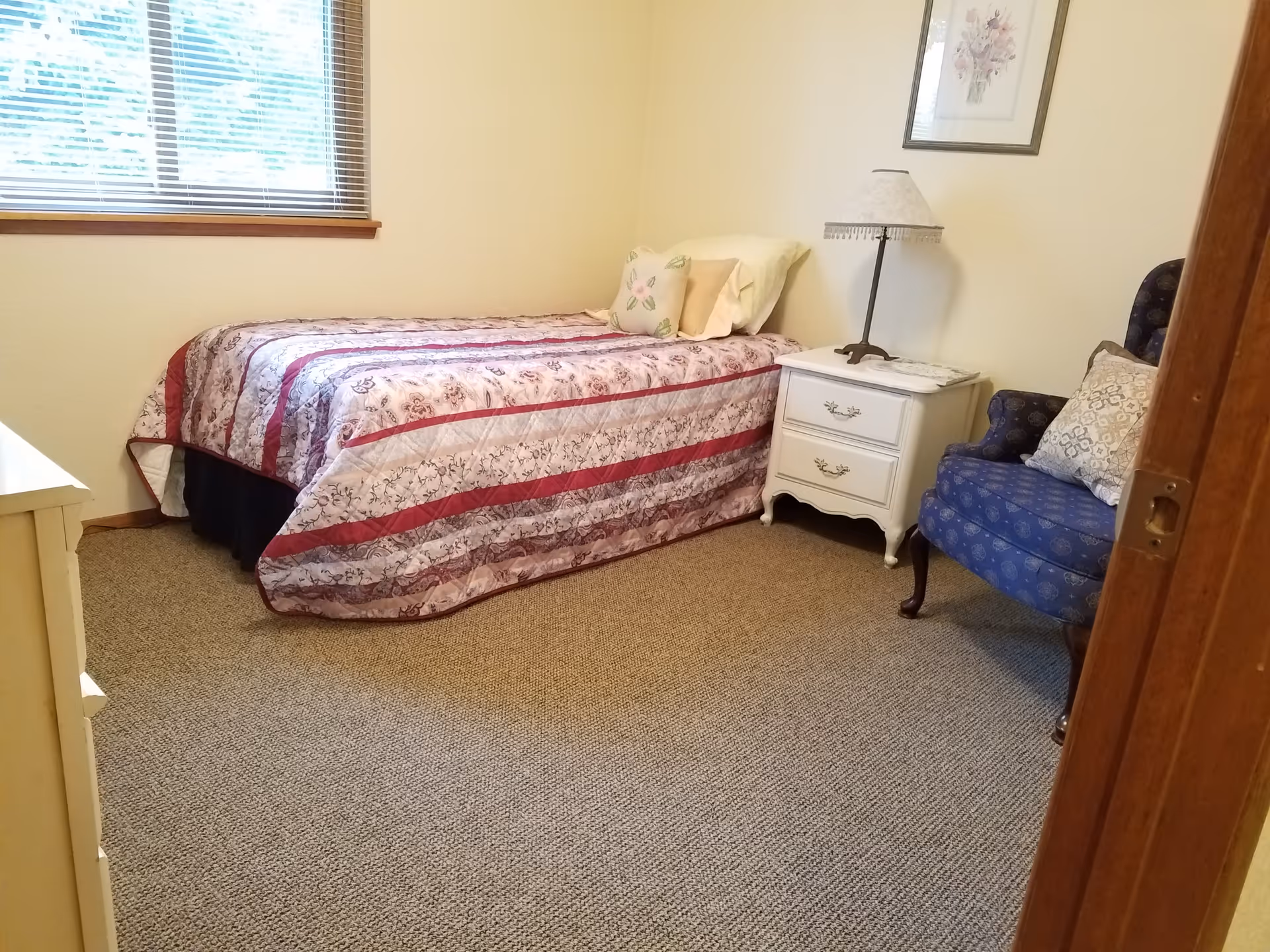 A neatly made single bed with quilt, nightstand, lamp, and blue chair in a cozy bedroom