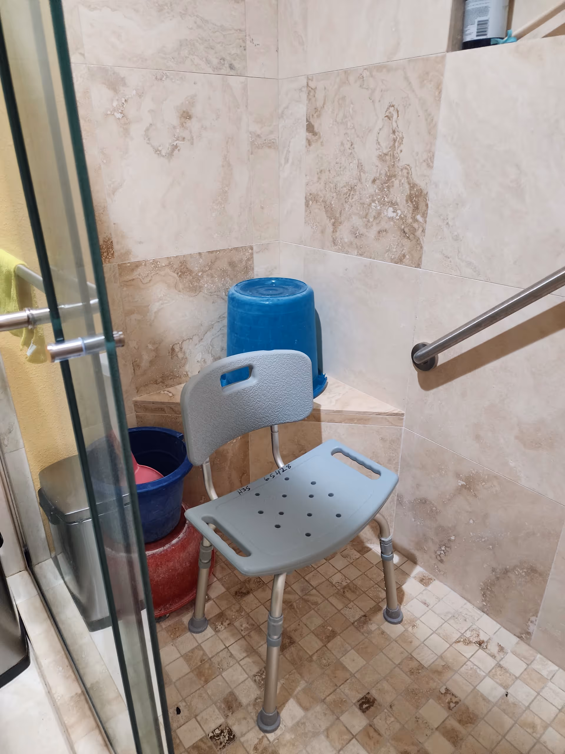 Small tiled bathroom with a glass shower enclosure, blue bucket, and folding chair.