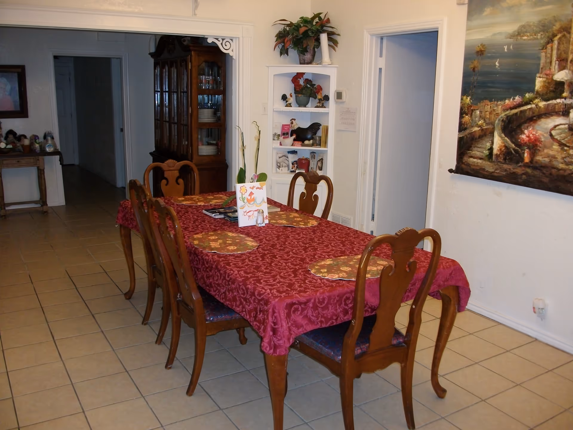 Dining room with a wooden table covered in a red tablecloth surrounded by chairs, a china cabinet, and wall art.