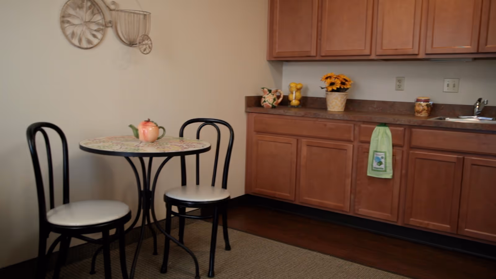 Small kitchen nook with a round bistro table and two chairs next to wooden cabinets and a countertop with a sink and decor.