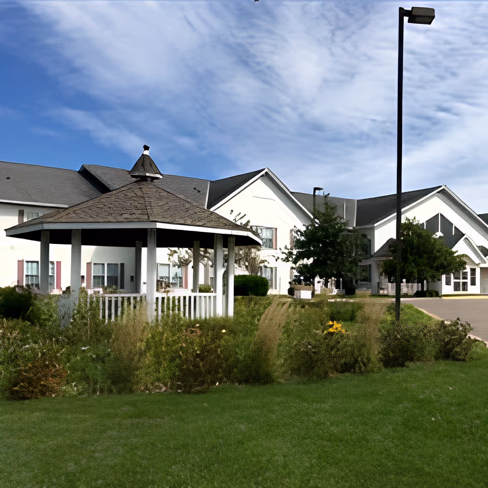 Outdoor view of Ecumen Hutchinson The Pines facility showing a white gazebo with a shingled roof surrounded by greenery and flowers, with the building and a tall street lamp in the background under a partly cloudy blue sky.