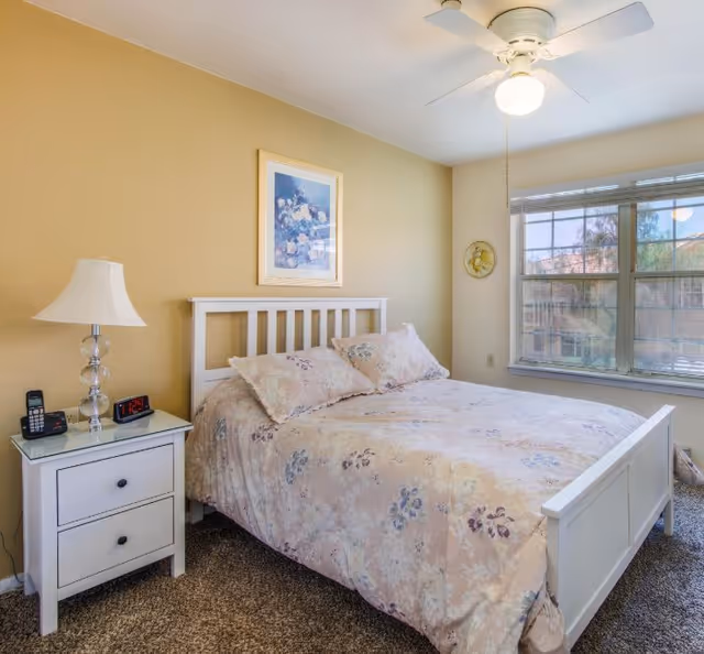 White bed with a floral quilt in a sunny bedroom, with a nightstand, lamp, and window.