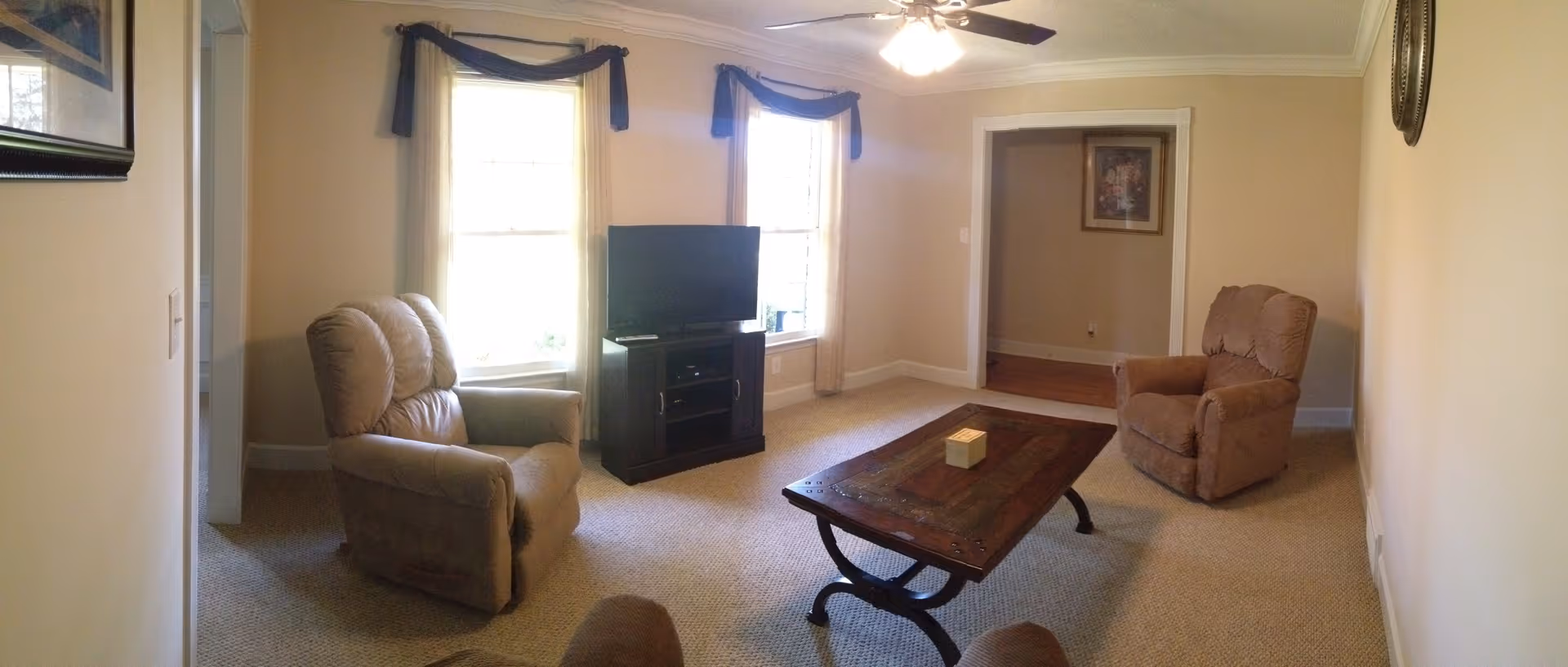 A cozy living room with beige walls and carpet, featuring two recliner chairs, a wooden coffee table with a small decorative item on it, a TV on a stand between two windows with dark valances, and a ceiling fan with lights.