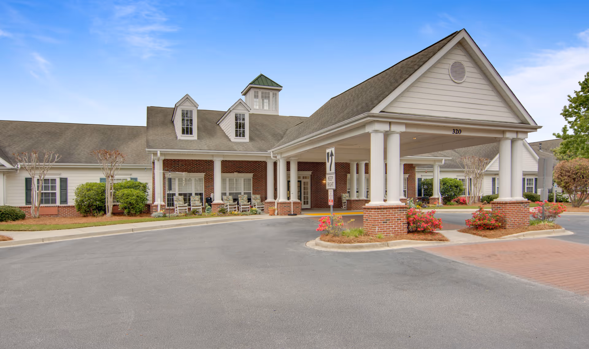 Front entrance and covered porte-cochere of a single-story residential building with white columns, brick accents, and landscaped beds.