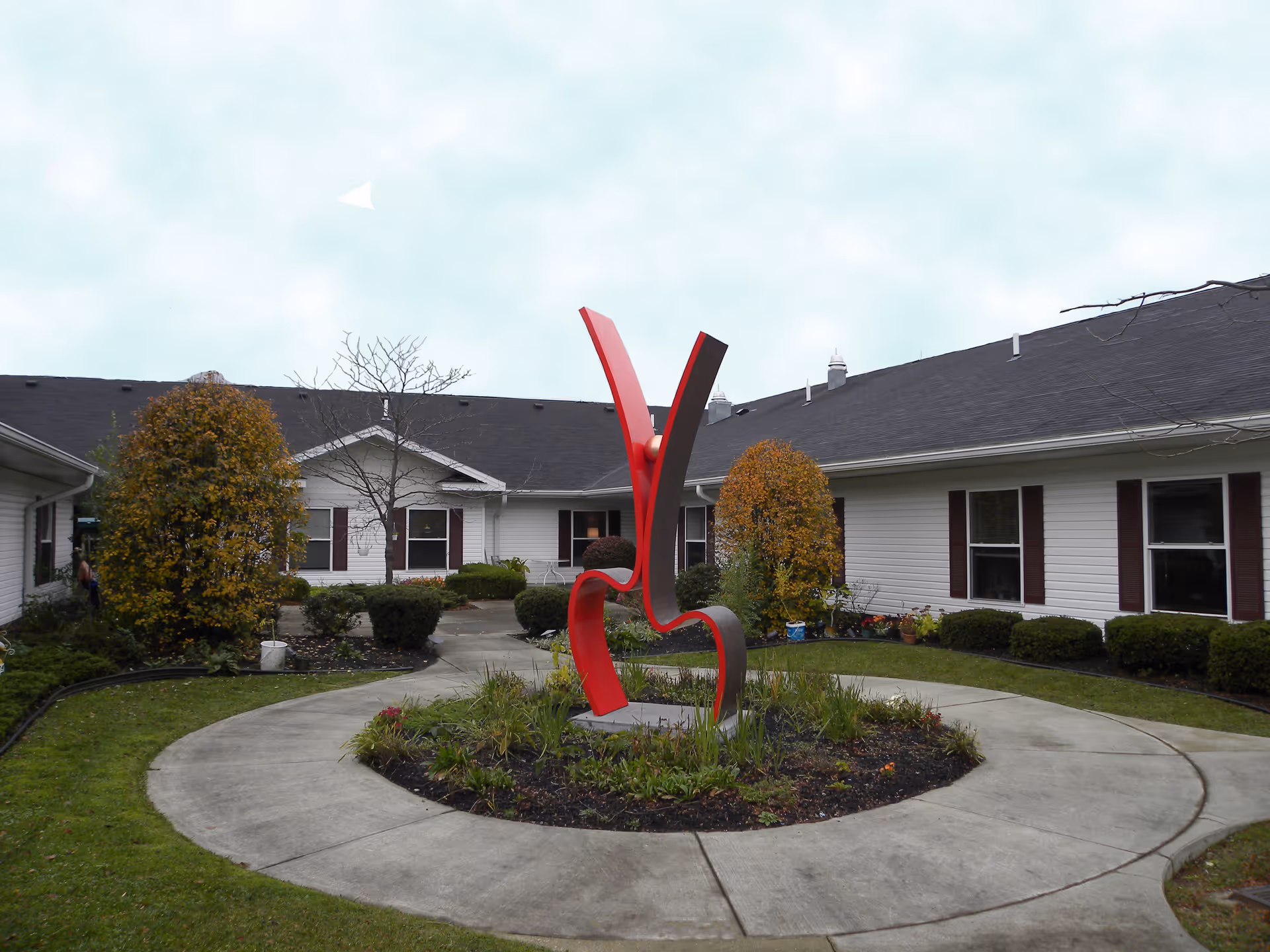 Courtyard of a senior living facility featuring a red abstract metal sculpture in the center surrounded by a circular walkway and single-story buildings.